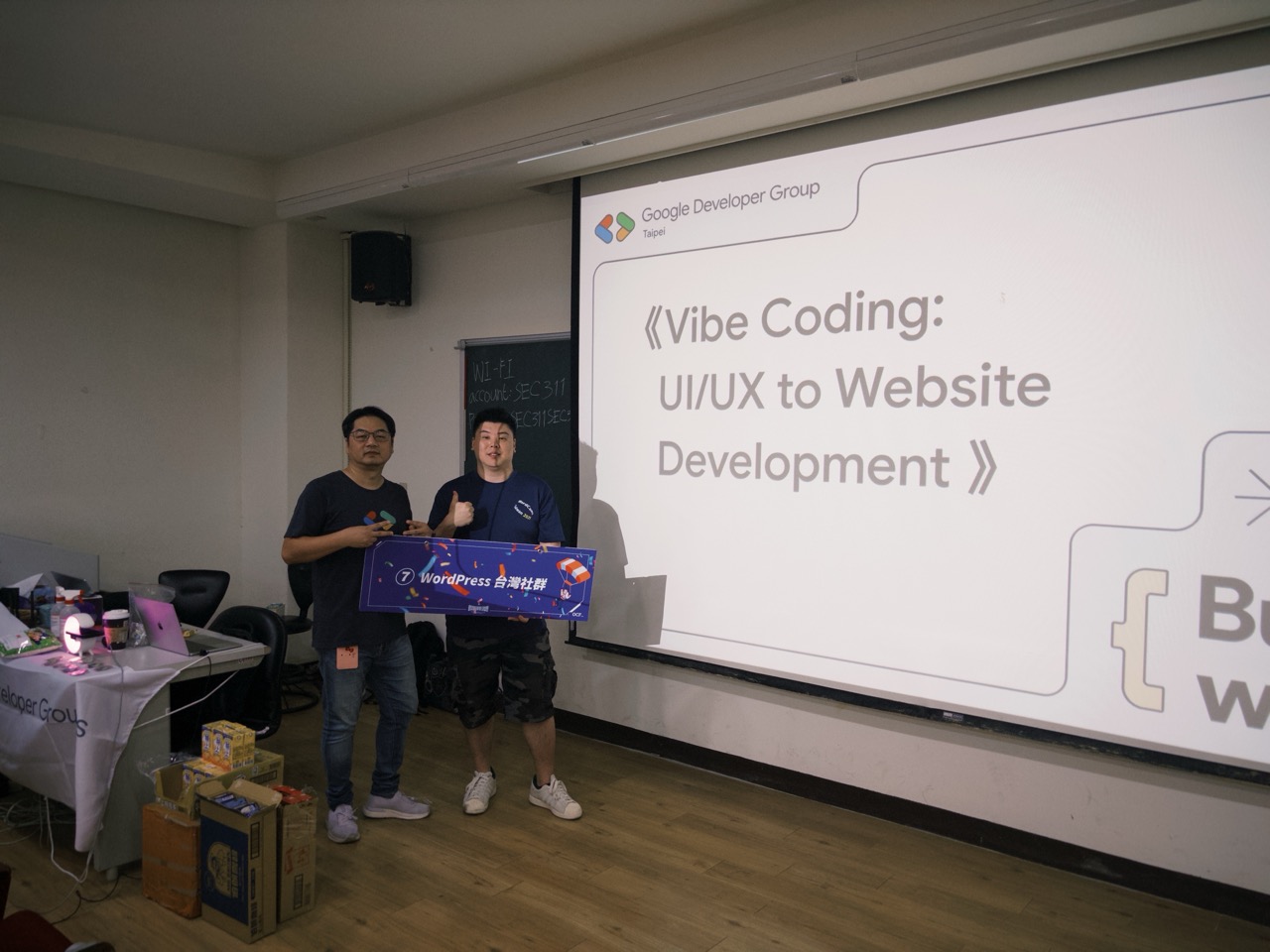 兩位主辦人拿著 WordPress 彩虹小聚旗幟，站在 Vibe Coding 簡報前合影