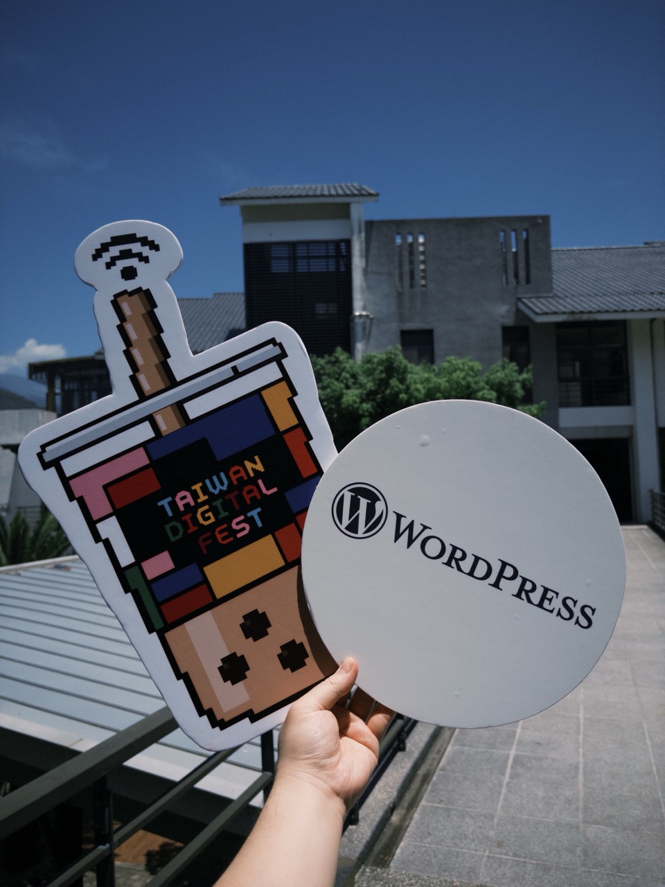 手持 Taiwan Digital Fest 與 WordPress 的造型扇，背景是臺東的校園建築