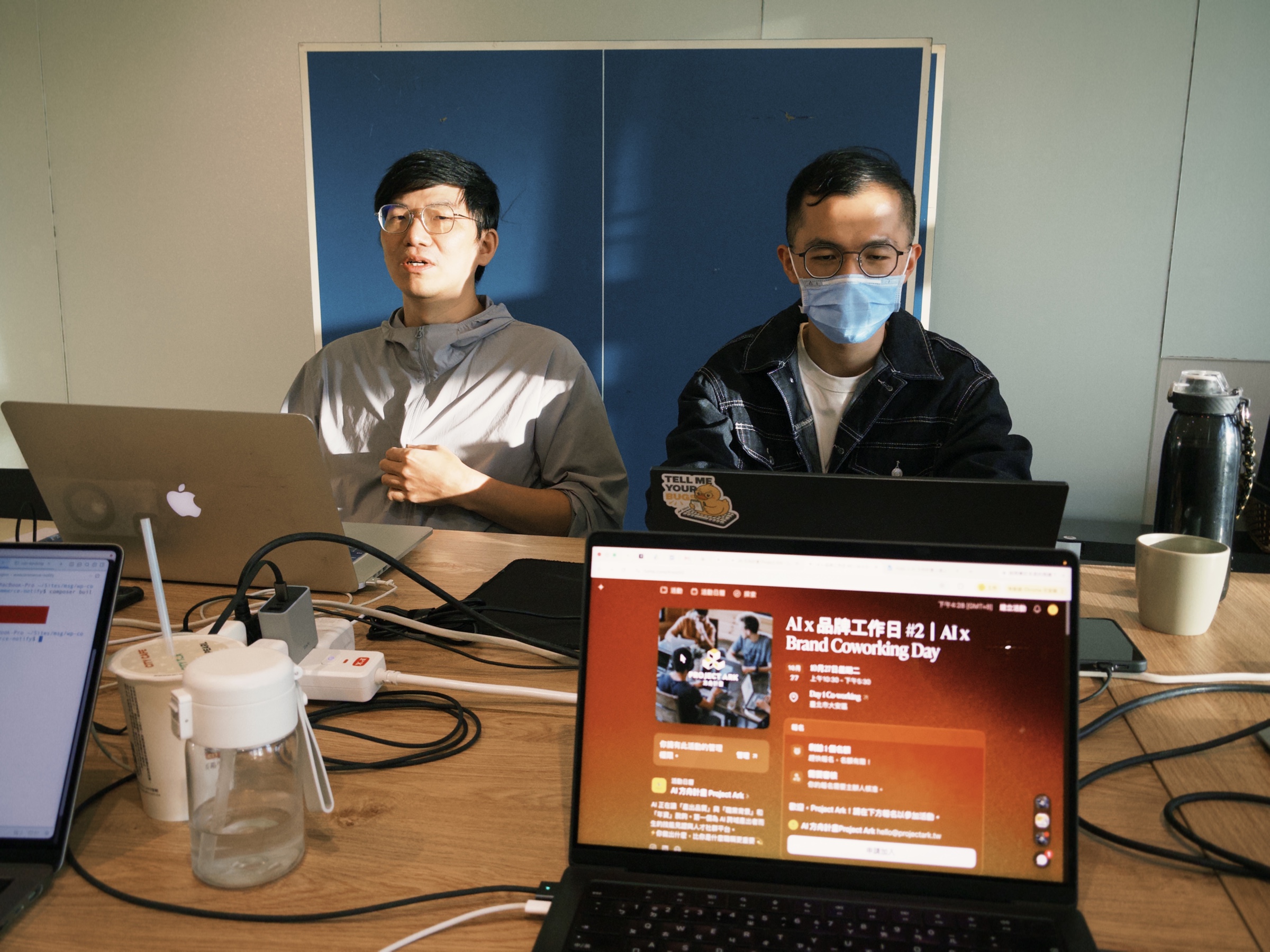 兩人並肩坐著交流，前方筆電螢幕顯示活動介紹頁面，桌上有飲料與水瓶